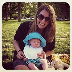 Person with long hair and sunglasses sitting on the grass, holding a baby wearing a bib and a blue hat